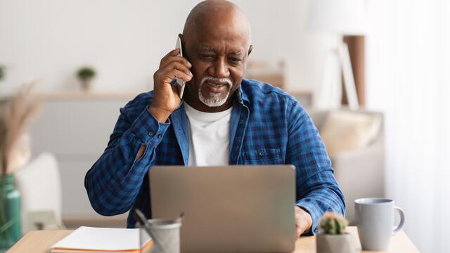 Adult talks on phone while sitting at desk and using a laptop