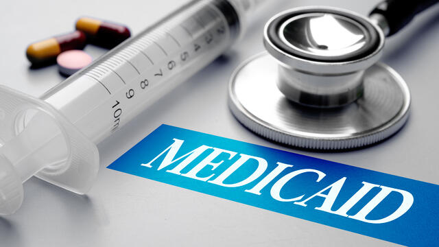 The word, Medicaid, is surrounded by brown pills, empty syringe and stethoscope