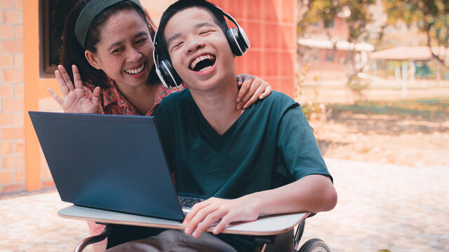 Child wearing headphones in a wheelchair with a laptop laughing with an adult