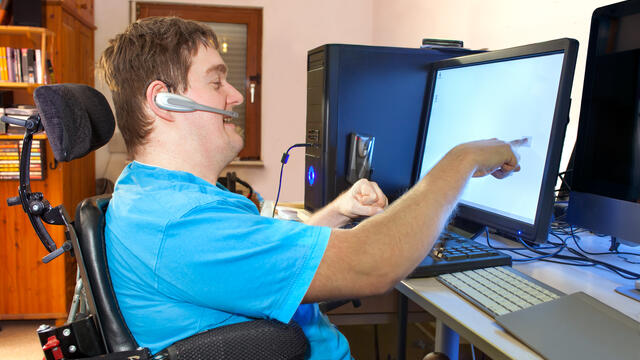 Adult in a wheelchair wears a headset uses a touch screen computer monitor