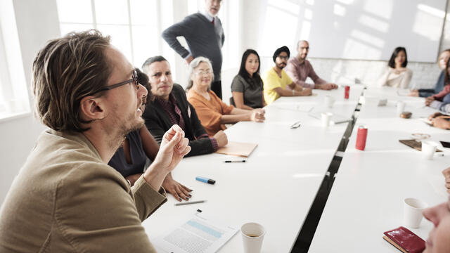 Adults sitting around a table and listening to a coworker during a meeting.