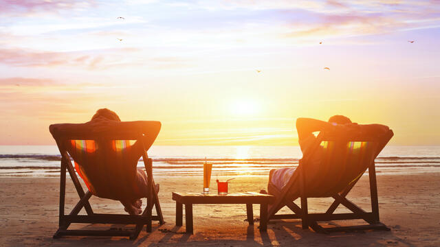 Silhouette of two adults in lawn chairs at a beach with drinks on a table.