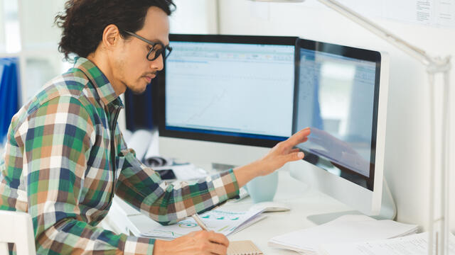An adult makes notes in a notebook at a desk with a computer monitor.