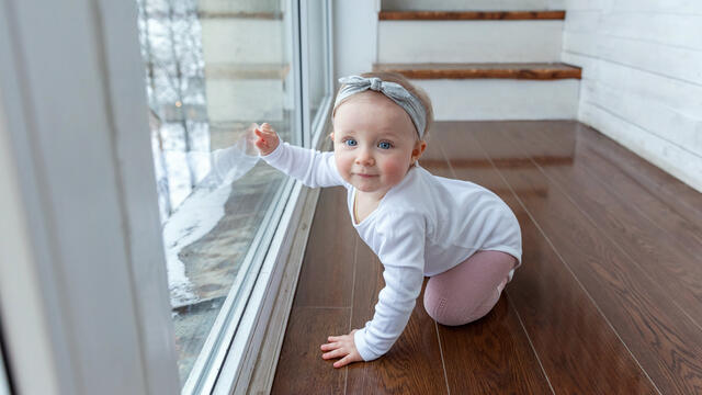 Baby on floor front of window with one hand on it.