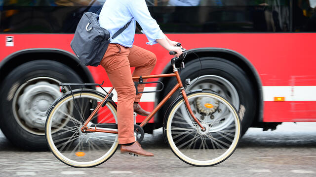 An adult with a briefcase rides bicycle next to a bus.