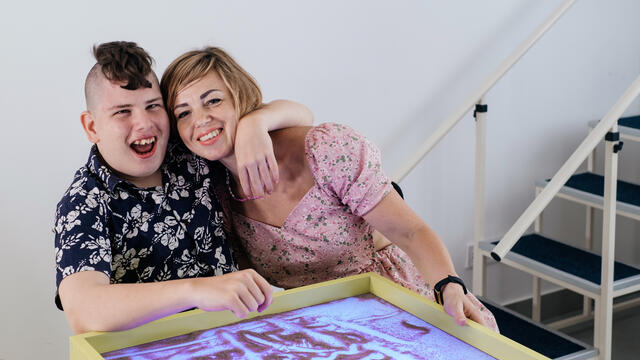 Two smiling adults with arms around each other in front of an art project