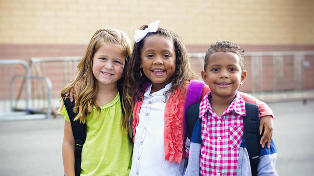 Three smiling children with arms around each other at a school.