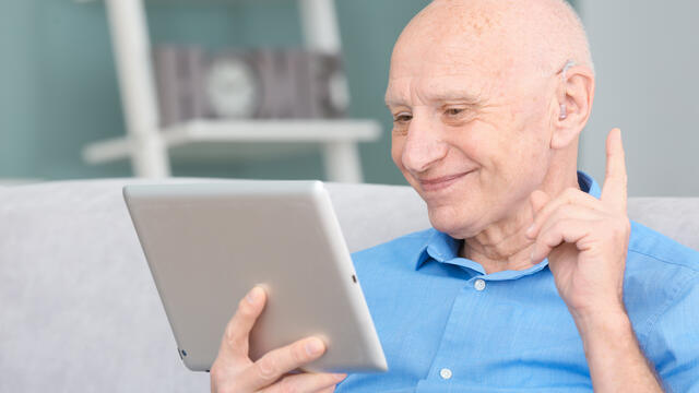 An older adult with a hearing aid smiles at a tablet