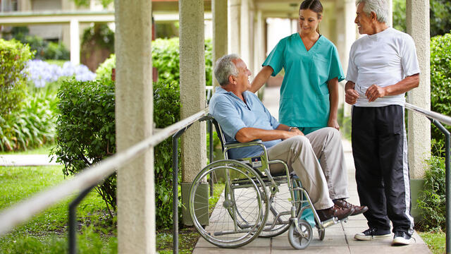 Caretaker talks to a person in a wheelchair at a living facility with another person standing by outside