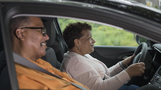 Two smiling adults sit in a care. Both wear seatbelts