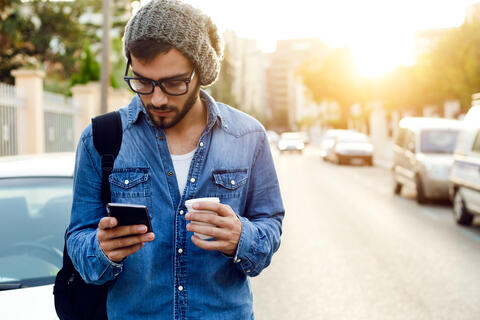Young adult with a cup walks in the street and checks his phone