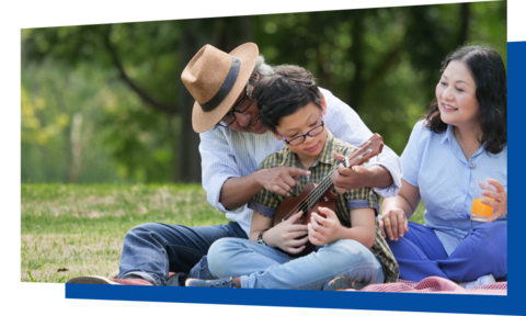 Adult holding glass watching another adult showing a child how to play a ukulele outdoors