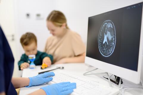 Parent and child meeting with a pediatric neurologist sitting at a desk with a brain scan on their computer