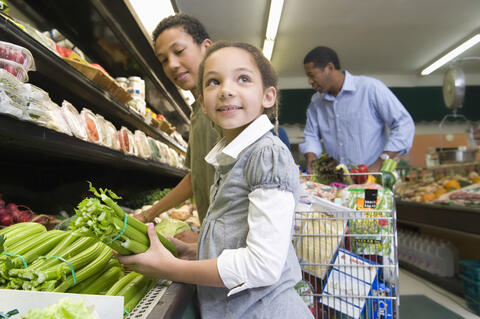 A child holding celery while adults look at vegetables in produce aisle.