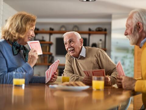 Three older adults playing cards in a home with snacks