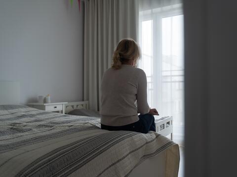 Back of a sad adult sitting on a bed looking out a window