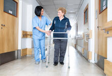 An adult assisting an older adult to walk down a hallway using a walker