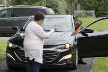 Person standing in driveway in front of a car with its driver's side door open. The person is giving the driver in the car a thumb's up gesture