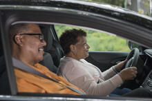 Two smiling adults sit in a care. Both wear seatbelts